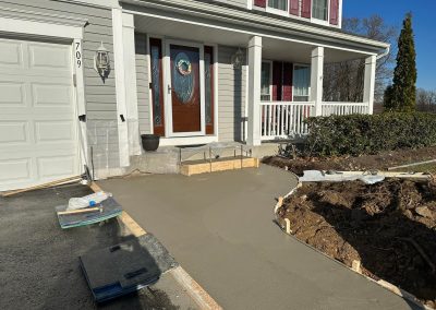 Freshly poured concrete sidewalk and steps in front of a house, with wooden forms and construction tools visible.