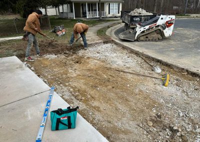 Two workers dig up a sidewalk area near a house; a skid steer and tools are visible nearby.