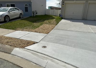 A concrete driveway and sidewalk beside a patchy lawn in front of two houses.