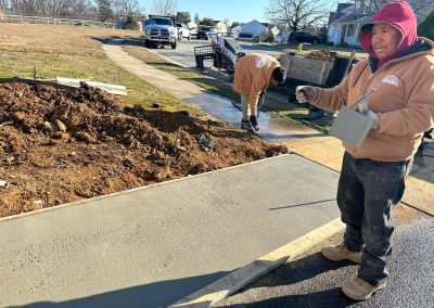 Two workers pour and smooth fresh concrete on a driveway near a suburban street on a sunny day.