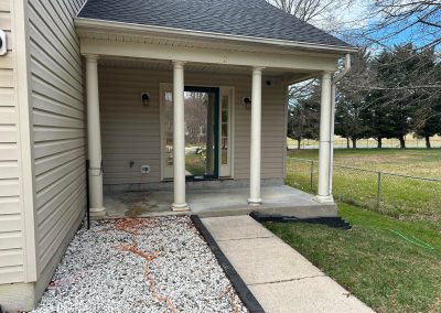 A beige house with a covered porch, four columns, gravel, and a concrete walkway leading to the front door.