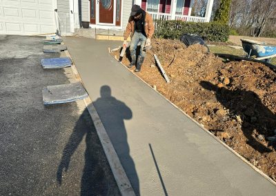 A worker smooths freshly poured concrete for a sidewalk outside a house on a sunny day.
