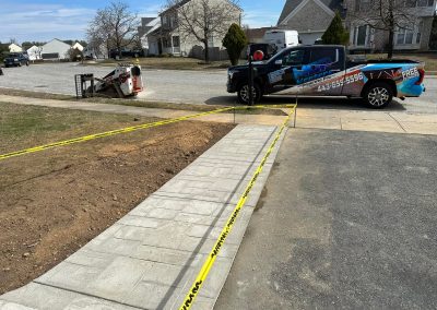 Newly paved sidewalk section bordered by caution tape, with a work truck and equipment nearby.