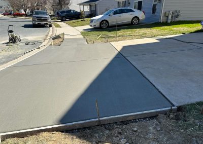 Freshly poured concrete driveway section next to older concrete, with cars and houses in the background.