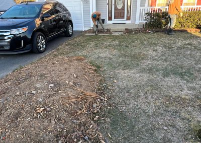 Two people work on a bare patch near the front door of a house, with a black car parked nearby.