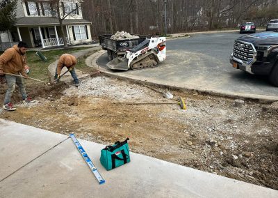 Two workers clear rubble from a driveway while a small loader and truck are parked nearby.