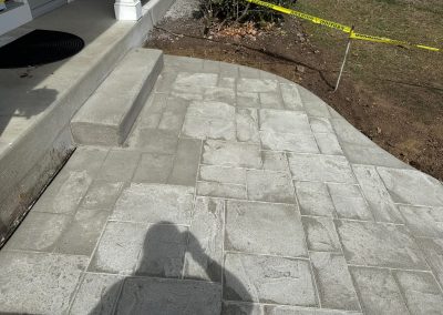Freshly laid concrete patio with caution tape and a step leading up to a porch; shadow visible in foreground.