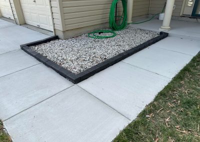 A gravel and stone garden bed bordered by bricks next to a garage, with a green garden hose coiled on top.