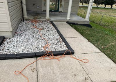 An orange extension cord stretched across a sidewalk next to a house with a gravel bed.