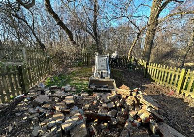 Broken bricks scattered before a toppled tombstone in a wooded, fenced area with trees and clear sky.