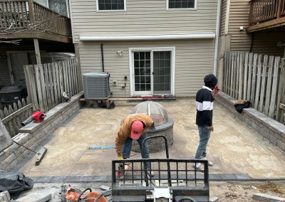 Two workers build a backyard patio with stone tiles and walls behind a house. Tools and equipment are visible.