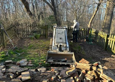 A person operates a small bulldozer near a fence, with bricks scattered in the foreground.