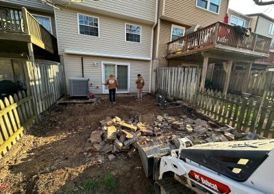 Two people stand in a backyard with broken concrete and debris, next to a small skid-steer loader and a fence.