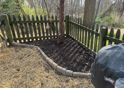 A fenced backyard corner with a mulched tree bed and a covered grill nearby on straw-covered ground.
