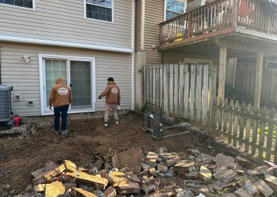 Two workers in brown hoodies prepare a backyard area for construction, with bricks piled in the foreground.