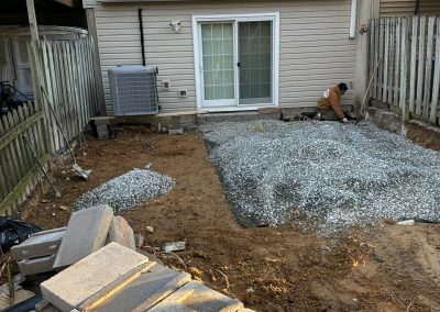 A person levels gravel in a backyard with stacked pavers and a sliding door on the house.