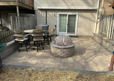A backyard patio with a round fire pit, outdoor dining set, and sliding glass doors to the house.