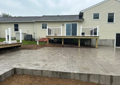 Freshly poured concrete patio and raised deck at the back of a light-colored house under a cloudy sky.