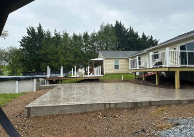 Freshly poured concrete slab in front of a house with decks and a pool, surrounded by greenery on a cloudy day.