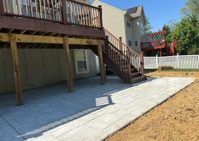 A backyard patio with stone pavers, a wooden deck above, and stairs leading down to the yard.