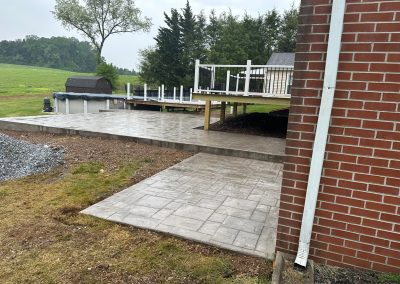A wet stamped concrete patio and walkway beside a brick house, with a raised deck and yard in the background.