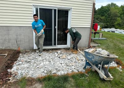 Two men break up and remove concrete near a house patio door; rubble is piled and a wheelbarrow is nearby.