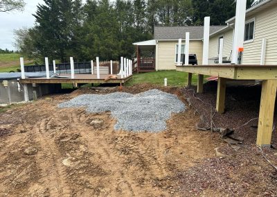 Construction site with dirt, gravel, and two partially built decks beside a beige house and trees in background.