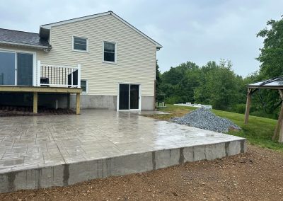 A newly poured concrete patio behind a beige house, with gravel and grass in the background.