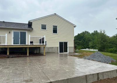 A beige house with a new deck, concrete patio, and a pile of gravel on a cloudy day.
