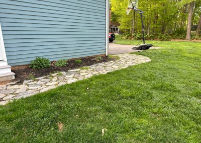 Stone path curves around a blue house with a basketball hoop and green lawn in the background.