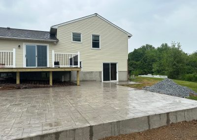 A beige house with a wooden deck and large concrete patio on a rainy day, surrounded by greenery.