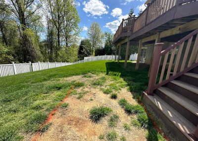 View of a backyard with patchy grass, red spray paint lines, white fence, and raised wooden deck stairs.