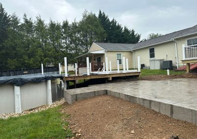 A backyard with a new wooden deck, above-ground pool, and concrete patio near a yellow house.