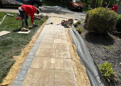 Two workers lay stone pavers on a walkway, with tools and landscaping materials nearby.