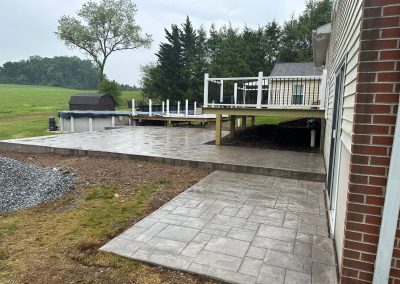 Gray stamped concrete patio and walkway beside a brick house, with a raised deck and pool in the background.