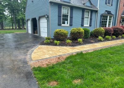 A stone walkway curves from a driveway toward a blue house with bushes along the path.