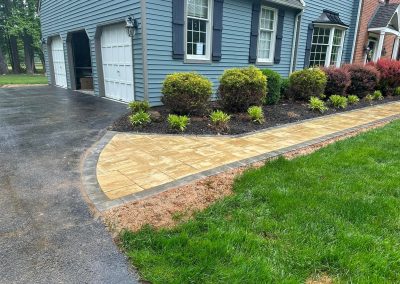 A paved stone walkway curves along a landscaped yard next to a blue house with a white garage.