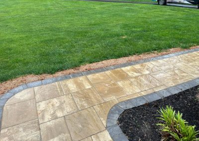 Wet stone path bordered with dark bricks, next to green grass and a small garden bed, with a car in background.