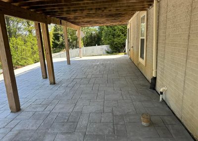 Covered patio with stamped concrete floor, wooden beams, and a beige brick wall next to a fenced yard.