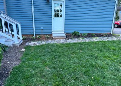 A white door on a blue house with stone path, green lawn, and small garden borders.