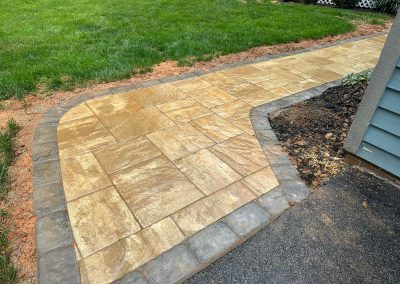 A wet stone walkway curves through grass and soil, bordered by gray pavers next to a building.