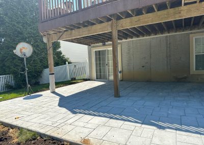 Concrete patio under a wooden deck with a satellite dish and white fence in a sunny backyard.