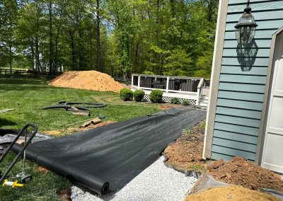 Black landscaping fabric is being laid beside a house with gravel and dirt piles nearby on a sunny day.