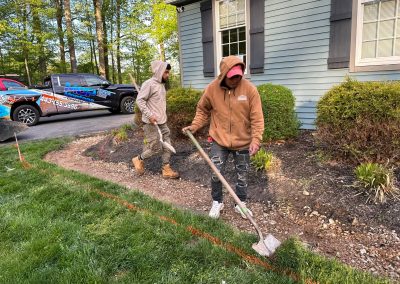 Two people in hoodies work with tools, preparing soil next to a house and bushes on a sunny day.
