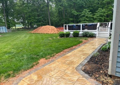A wet stone walkway leads to a deck and pool, with a dirt pile and grassy yard in the background.