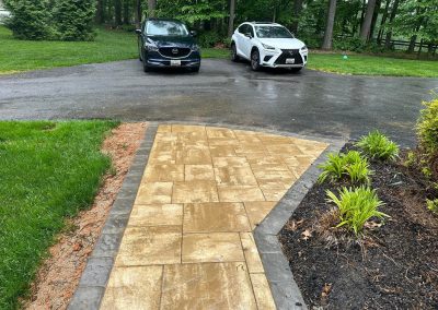 Yellow stone walkway leads to a driveway with two parked cars, surrounded by grass and trees.