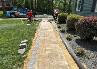 Workers install a stone walkway next to a house with grass, shrubs, and work vehicles in the background.