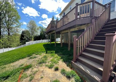Backyard with green grass, a brown raised deck, stairs, white fence, and a patch of dry grass below.