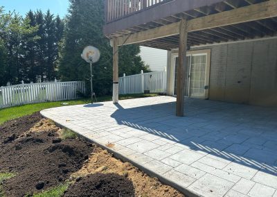 A newly paved patio under a wooden deck, beside a garden area with fresh soil in a fenced backyard.