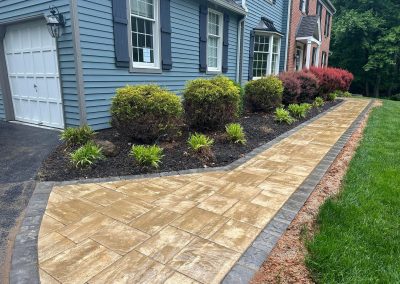 Tan stone walkway with a gray border runs beside a house, lined with shrubs and fresh mulch.